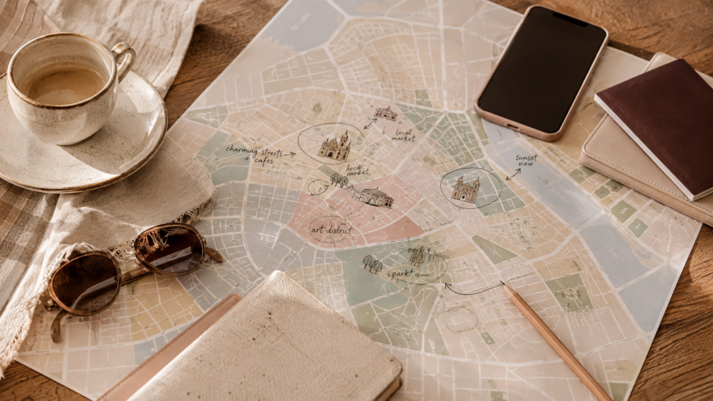 Elegant travel map spread across a warm wooden table with handwritten route markings, sunglasses, smartphone, coffee cup, and travel essentials.